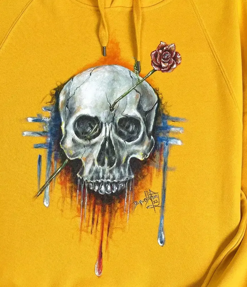 Close-up of a hand-painted skull and rose on a yellow hoodie, with the artist’s signature